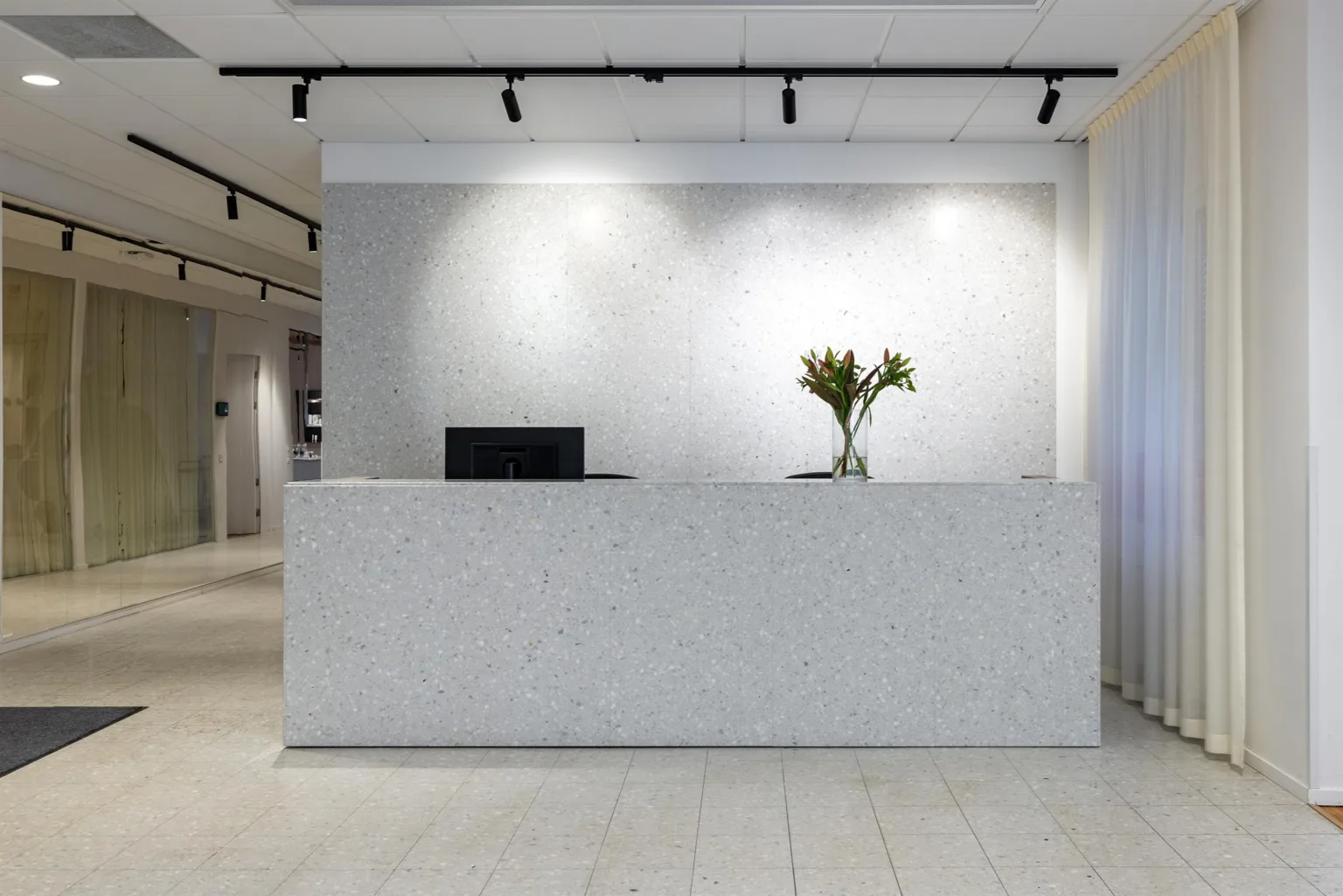 3. En reception med en vit disk, en datorskärm och en vas med blommor. Väggarna är vita och spotlights lyser upp området.