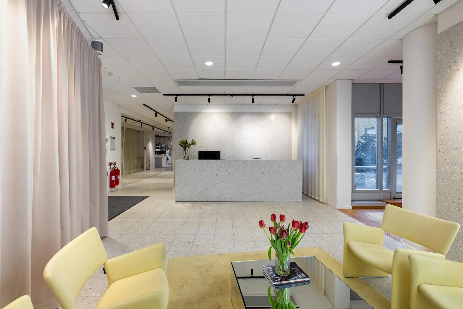 En reception med vit disk, gula stolar, ett glasbord med röda tulpaner och ljusa väggar.