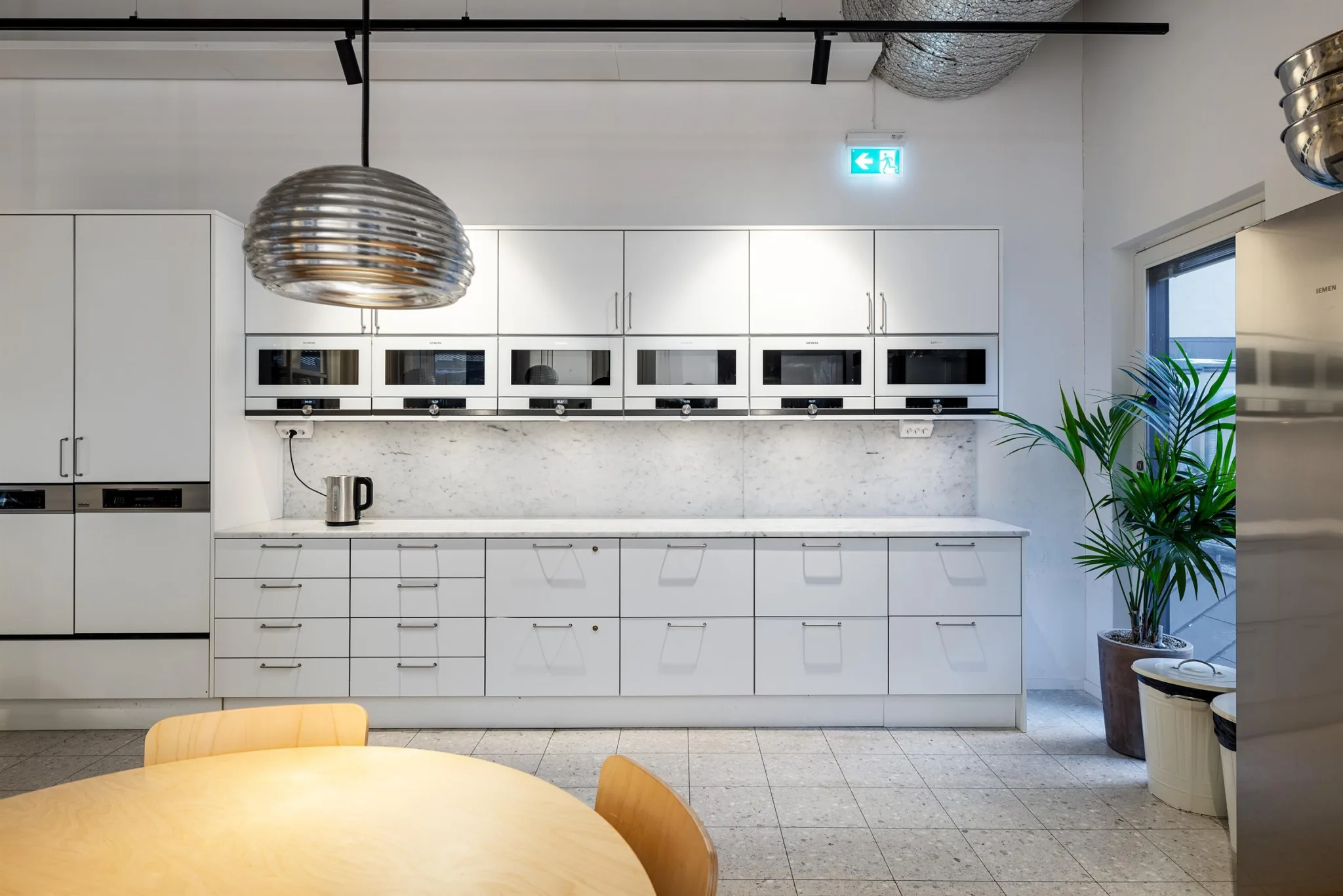 2. En ljus köksyta med flera inbyggda mikrovågsugnar, vita skåp och en rund matplats. En stor metallisk lampa hänger ovanför bordet.