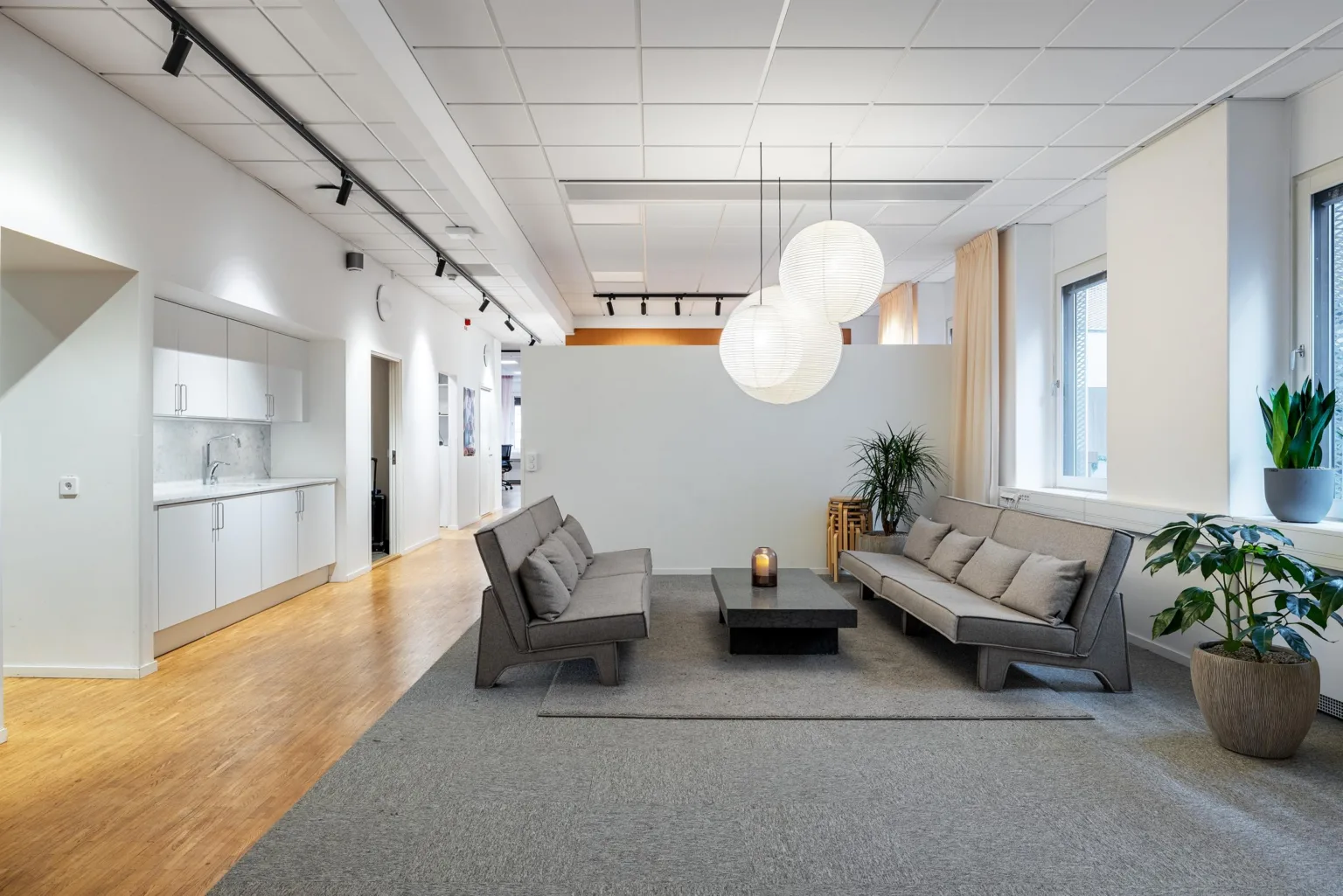 Ett modernt loungeområde med grå soffor, ett svart soffbord och stora vita taklampor, omgiven av ljusa väggar och fönster.