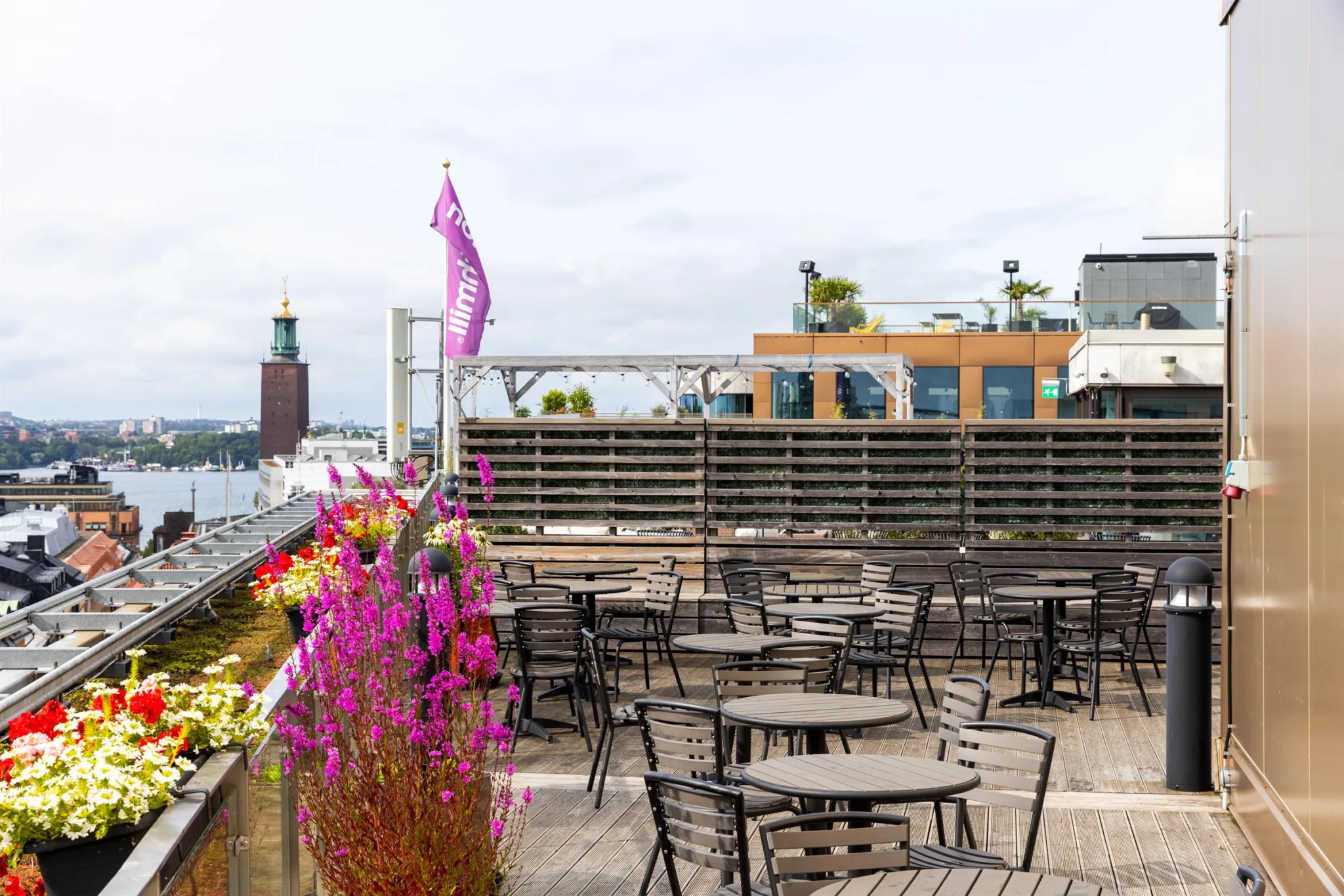En takterrass med flera bord och stolar, färgglada blommor och utsikt över stadens byggnader.