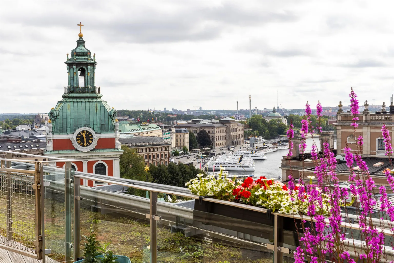 Utsikt över en stad med kyrka, vatten och blommor i förgrunden på en balkong.