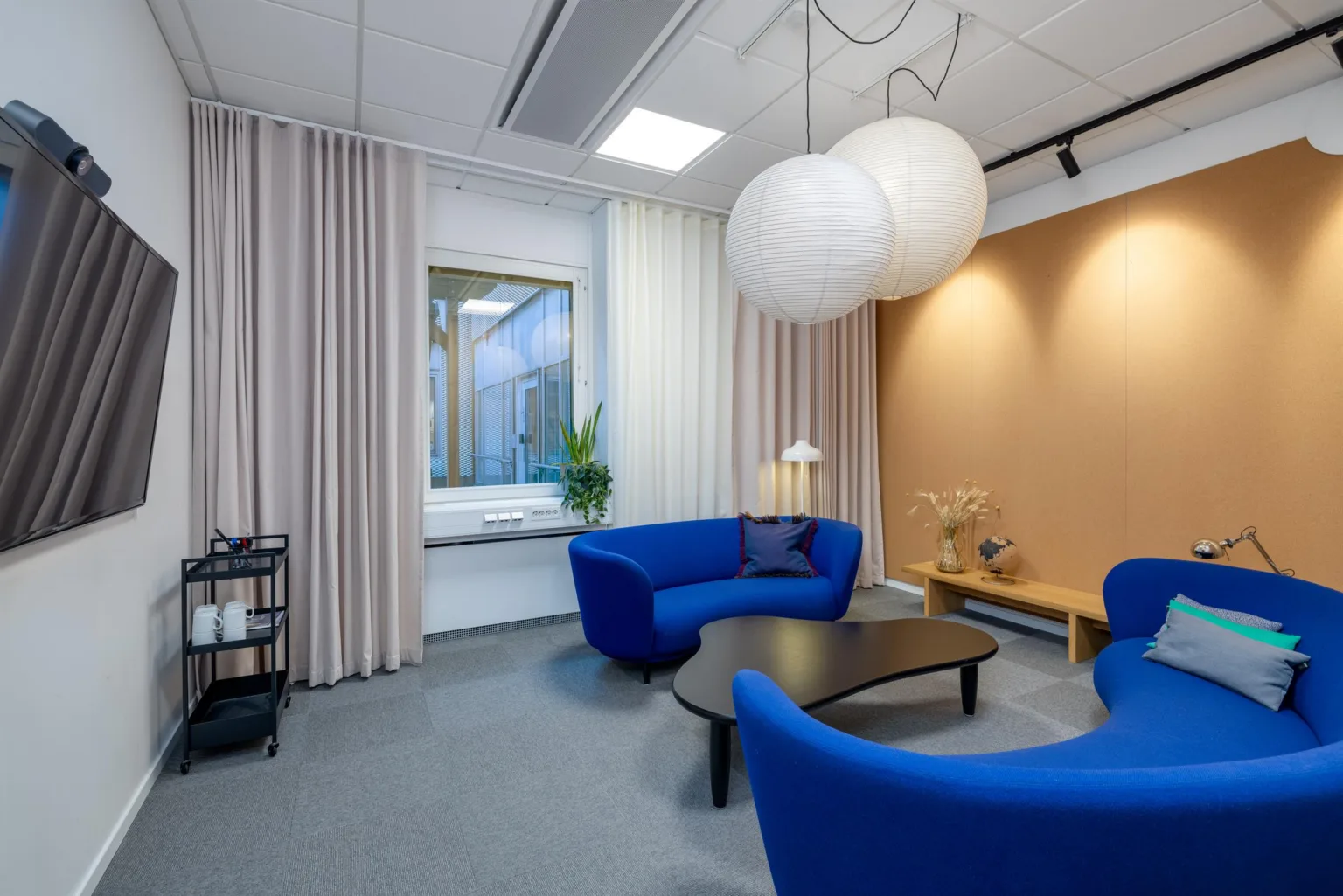 Ett litet loungeområde med blå soffor, ett svart bord, en stor vit taklampa och vita gardiner framför ett fönster.