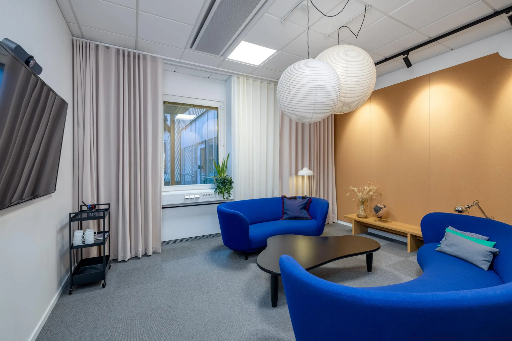 Ett litet loungeområde med blå soffor, ett svart bord, en stor vit taklampa och vita gardiner framför ett fönster.