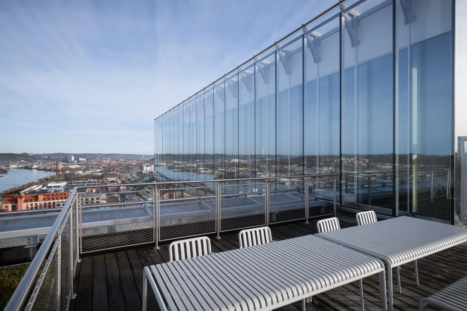 En takterrass med vita bord och stolar, omgiven av glasräcken med utsikt över staden.