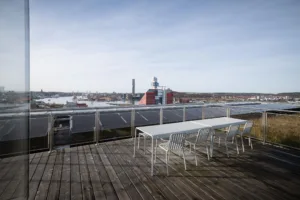 En takterrass med trämöbler och utsikt över en stad med industriella byggnader och vatten.