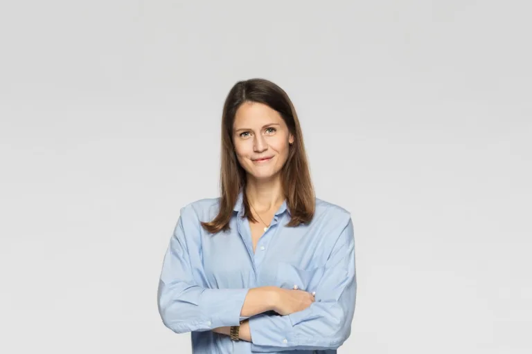 Kvinna med långt brunt hår och blå skjorta som står med armarna i kors och ler.