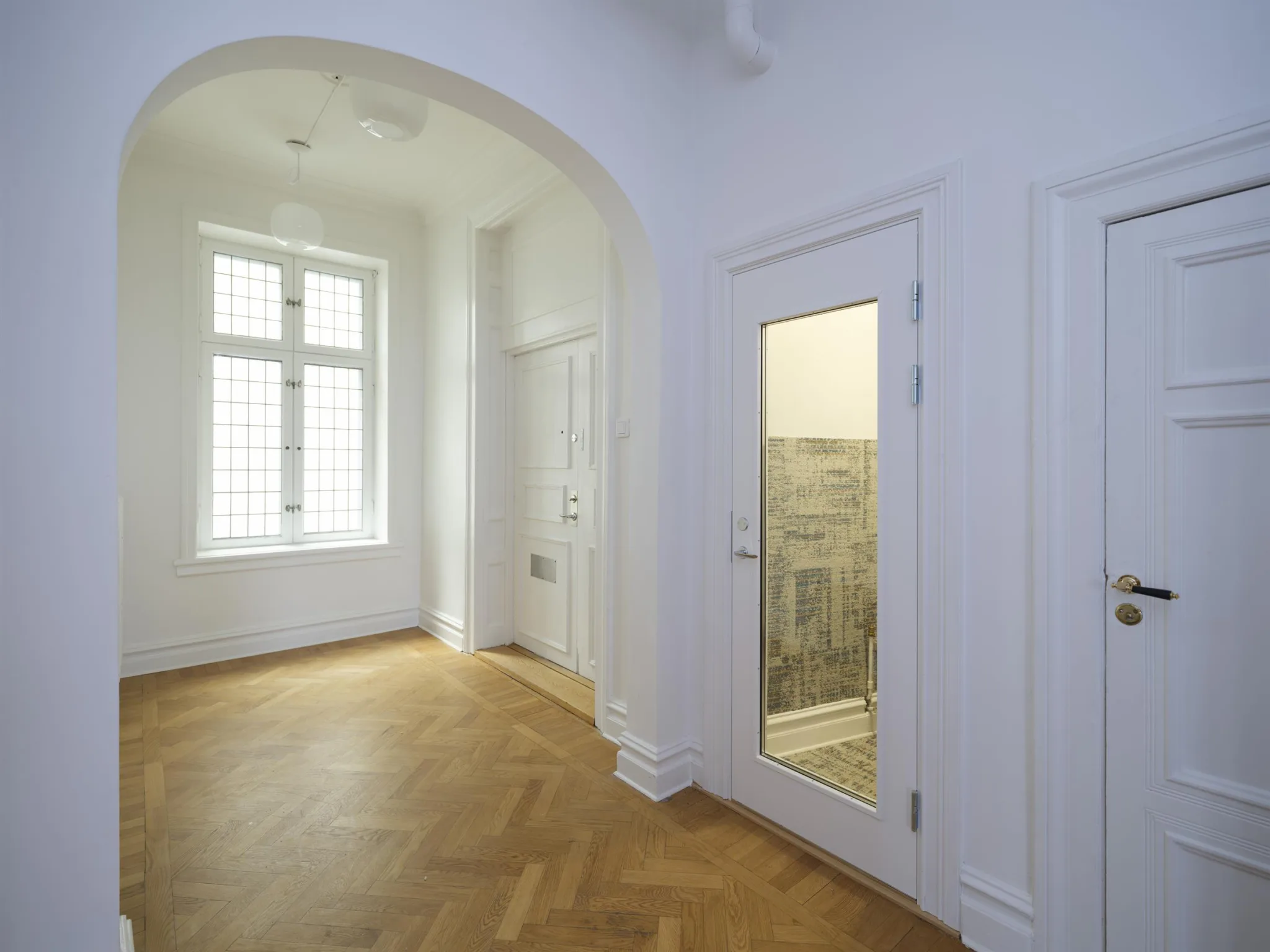 En hall med vita väggar, parkettgolv och en dörr med glas som visar ett kaklat badrum; fönster med ljusinsläpp finns i bakgrunden.