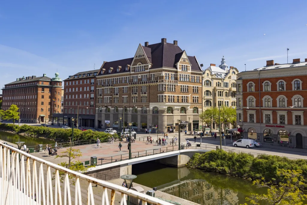 En stadsvy med klassiska byggnader, en bro och människor som promenerar vid en kanal under en klar blå himmel.