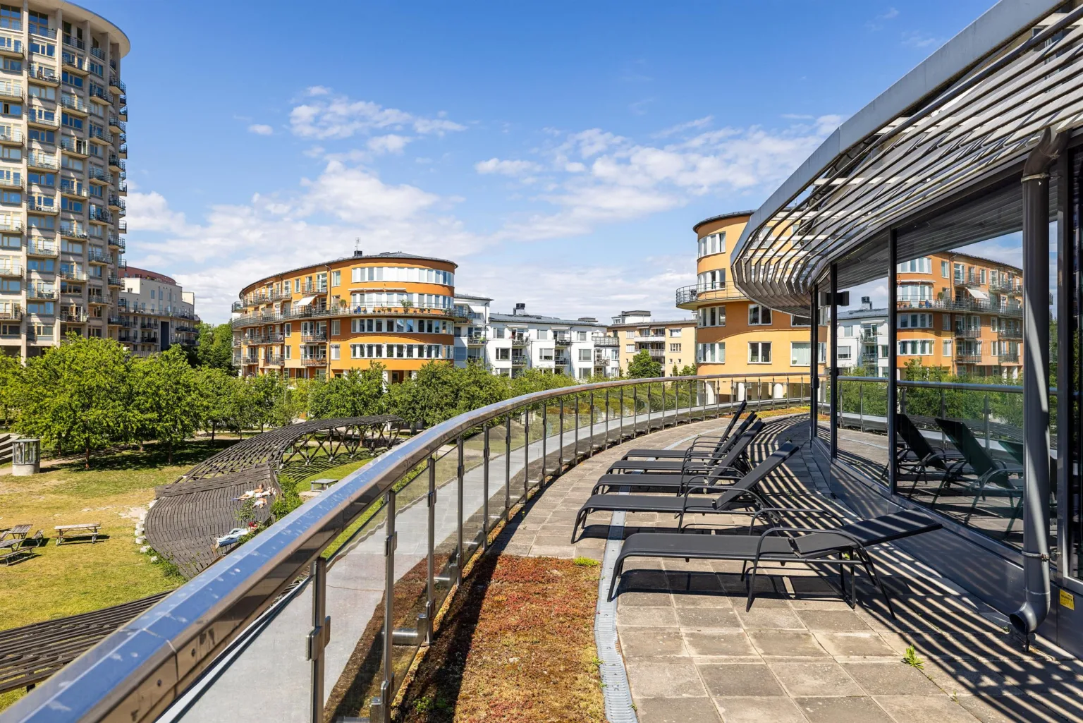 En terrass med glasräcke, solstolar och utsikt över en park med gröna omgivningar och flerbostadshus i bakgrunden.