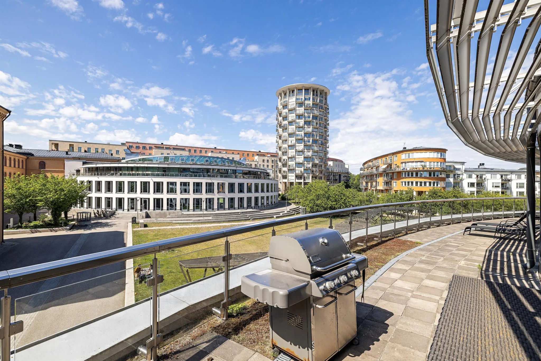 Balkong med grill, utsikt över en modern byggnad med rund fasad och grönska under en blå himmel.