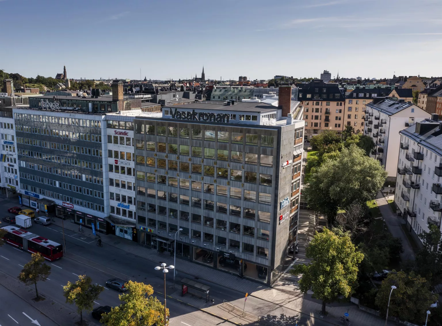 En kontorsbyggnad i flera våningar med glasfasad, omgiven av träd och stadsmiljö under en klar himmel.