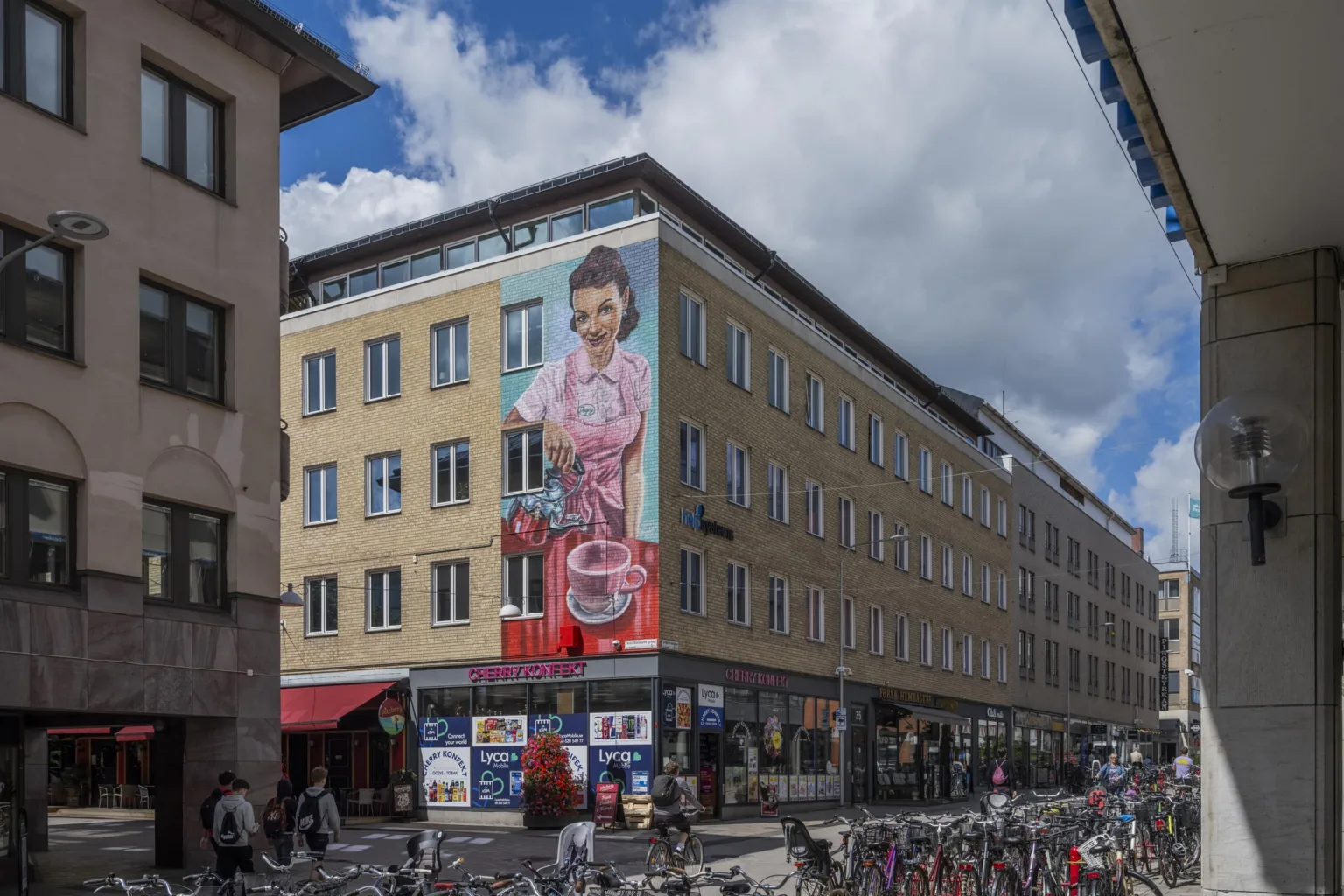 En äldre flerfamiljsbyggnad med en stor väggmålning föreställande en person ovanför affärer, cyklar parkerade längs trottoaren.