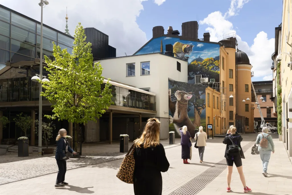 En gata med människor som promenerar, omgiven av byggnader. En stor muralmålning pryder en fasad i bakgrunden.