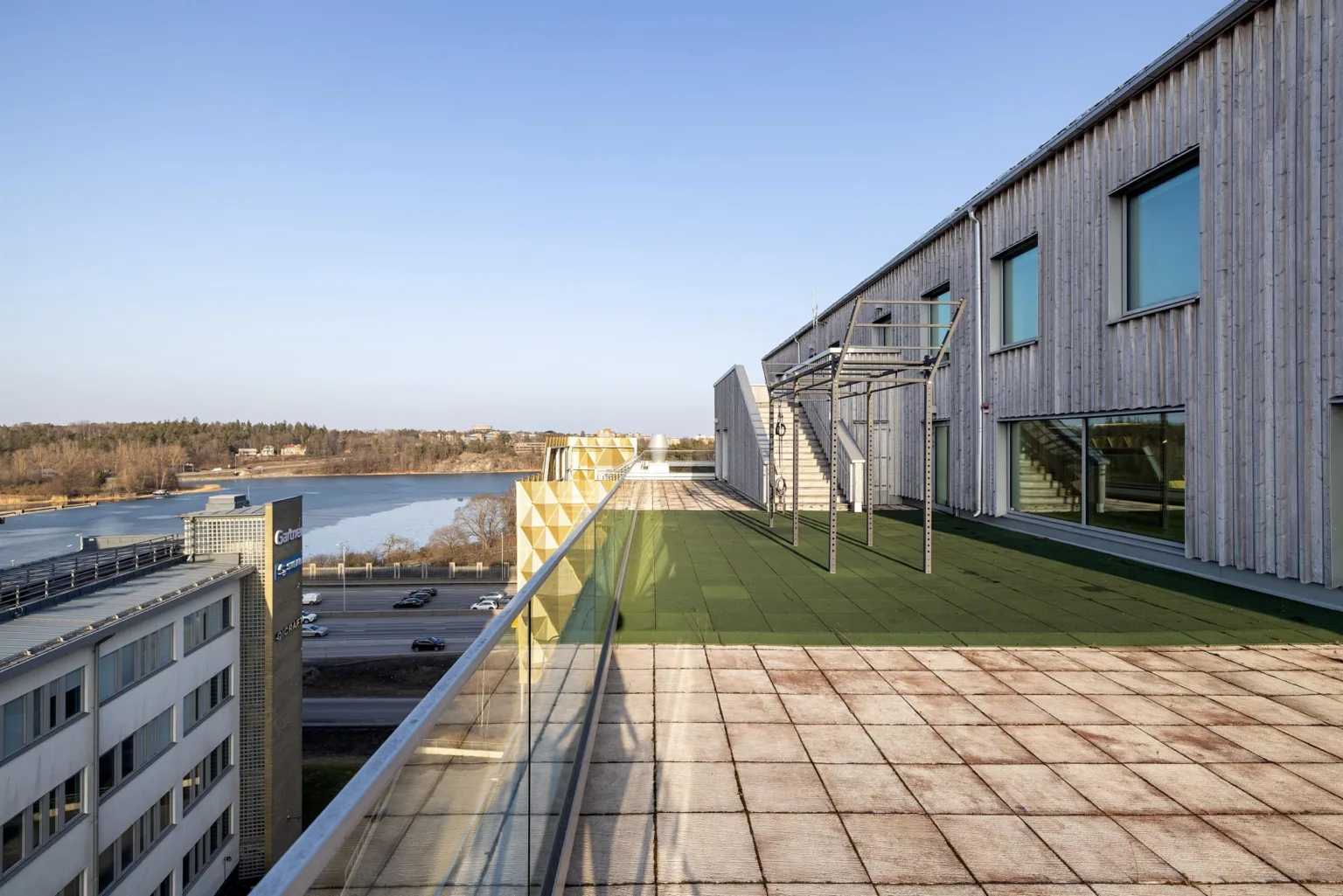 En takterrass med konstgräs och metallställningar, omgiven av glasräcken och utsikt över en sjö och byggnader.