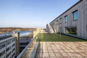 En takterrass med konstgräs och metallställningar, omgiven av glasräcken och utsikt över en sjö och byggnader.