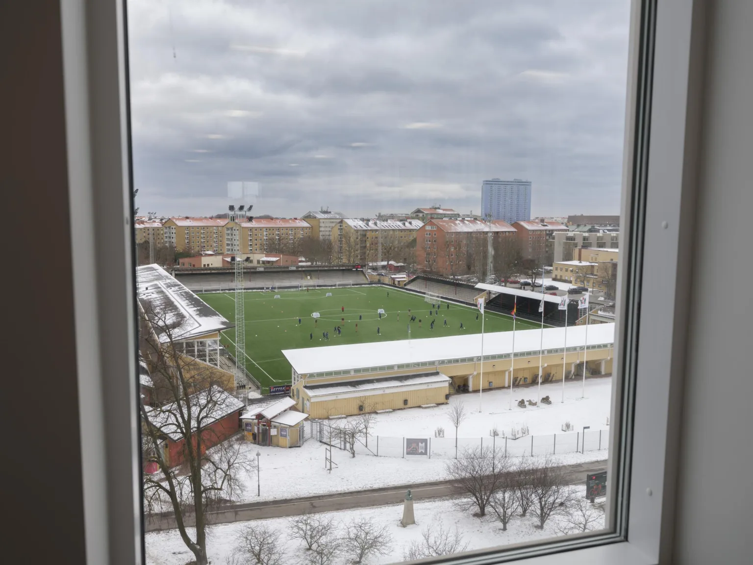 Utsikt från ett fönster över en snötäckt idrottsplan och omgivande byggnader.