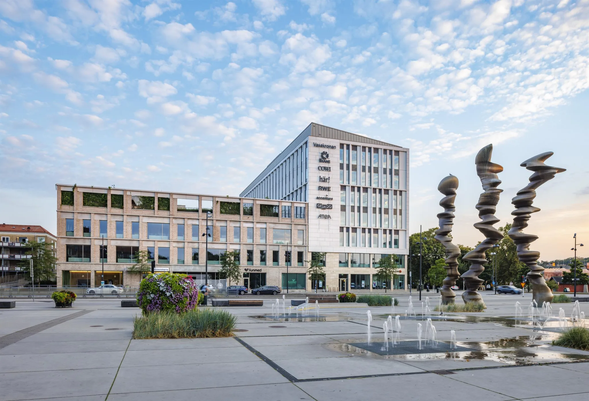 Ett torg med fontäner, en modern byggnad med glasfasad och en skulptur i förgrunden under en blå himmel med moln.