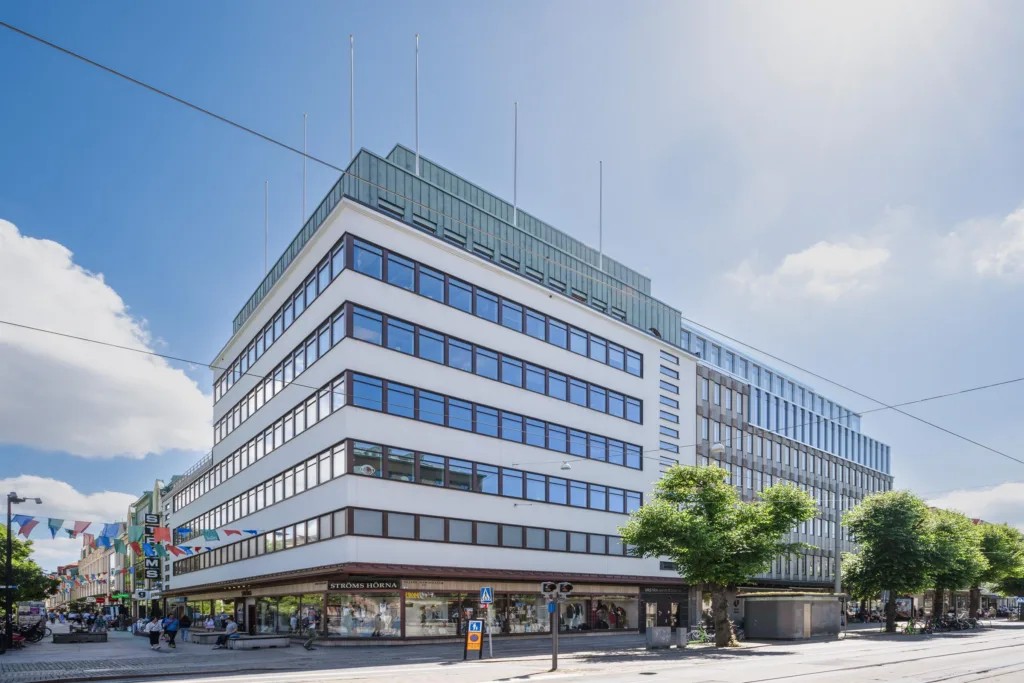 Modern vit byggnad med flera våningar, stora fönster och ett träd vid ingången.
