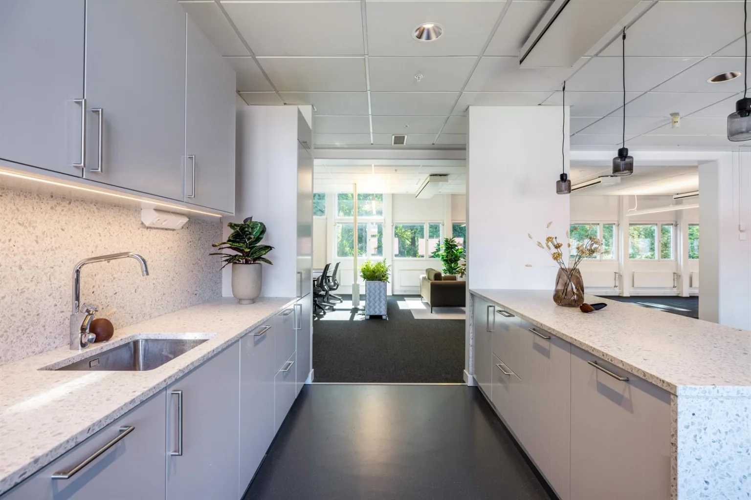 Ett modernt kök med vita skåp, två diskhoar, belysning och utsikt mot ett kontorsutrymme i bakgrunden.