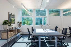 Ett ljust kontorsrum med två skrivbord, kontorsstolar och stora fönster som vetter mot grönska.