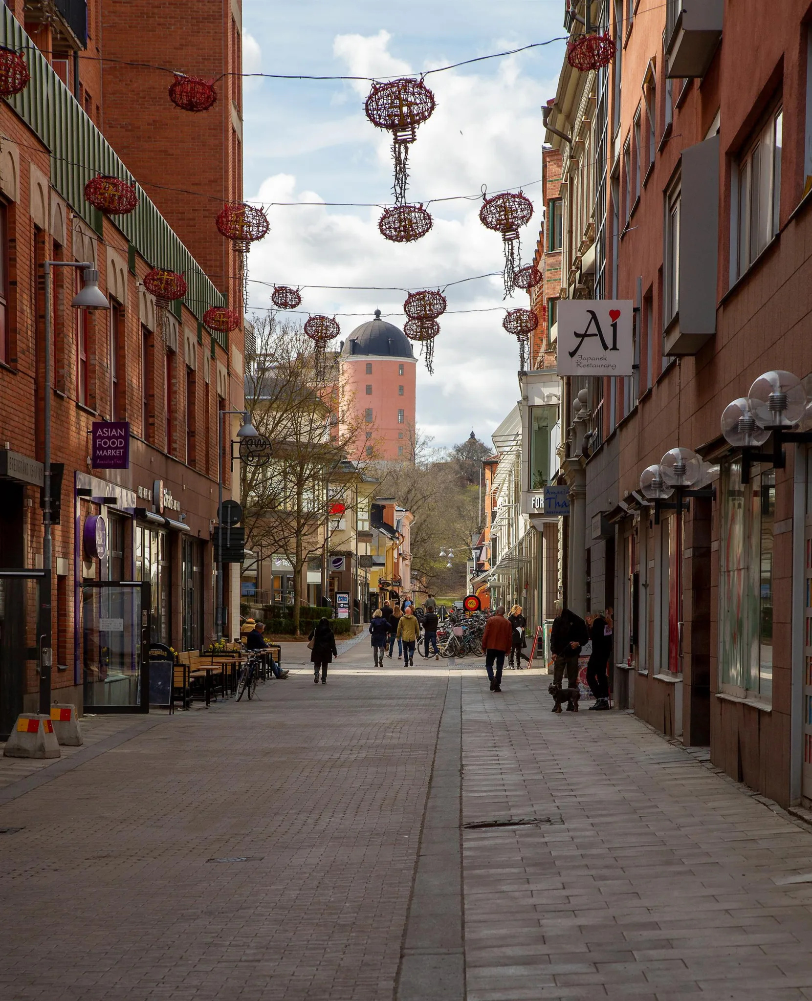 En gågata med röda lyktor hängande ovanför, omgiven av byggnader och människor som promenerar.