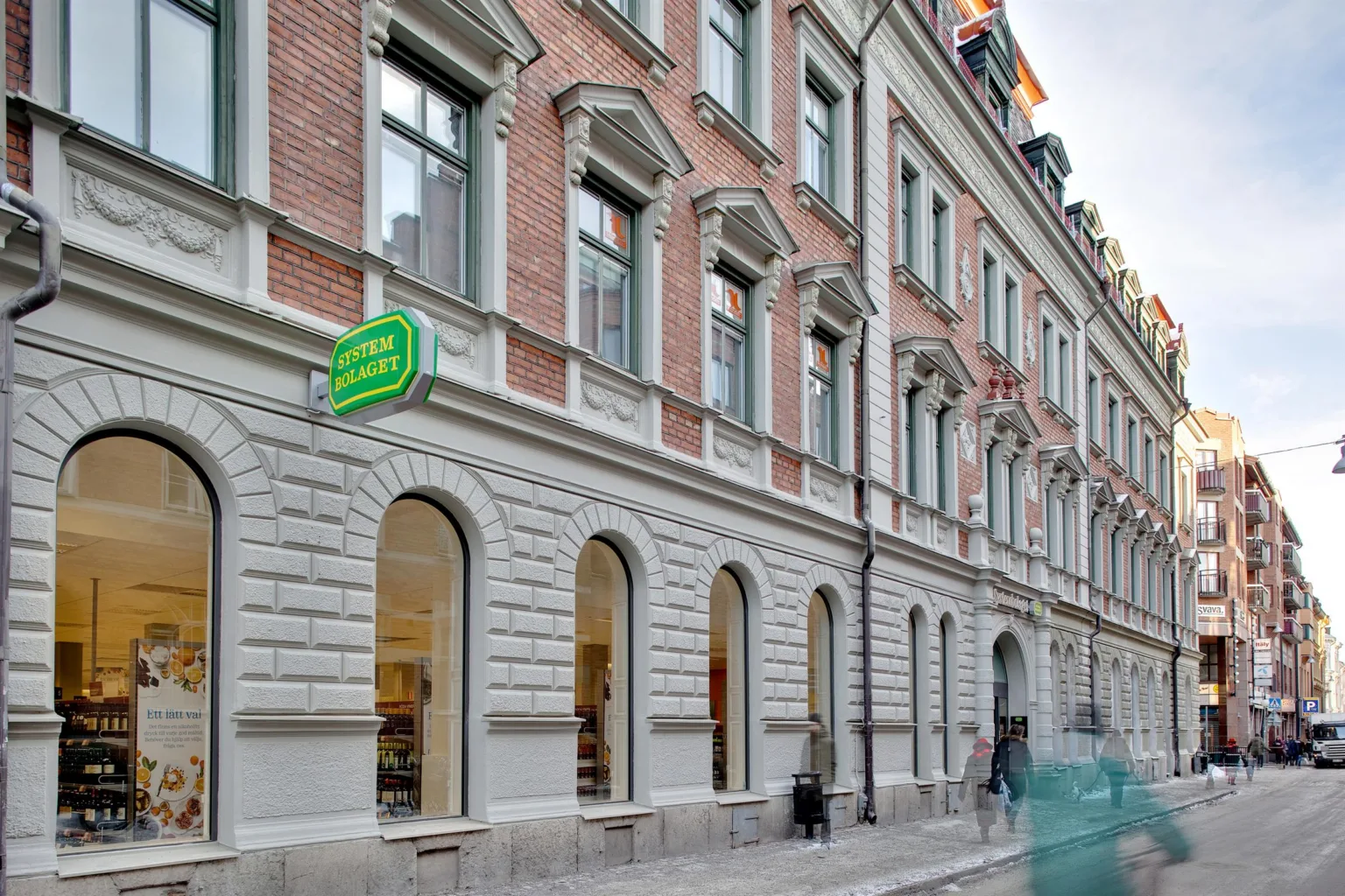 En fasad av en äldre byggnad med stora fönster och en grön skylt med texten "Hemköp".