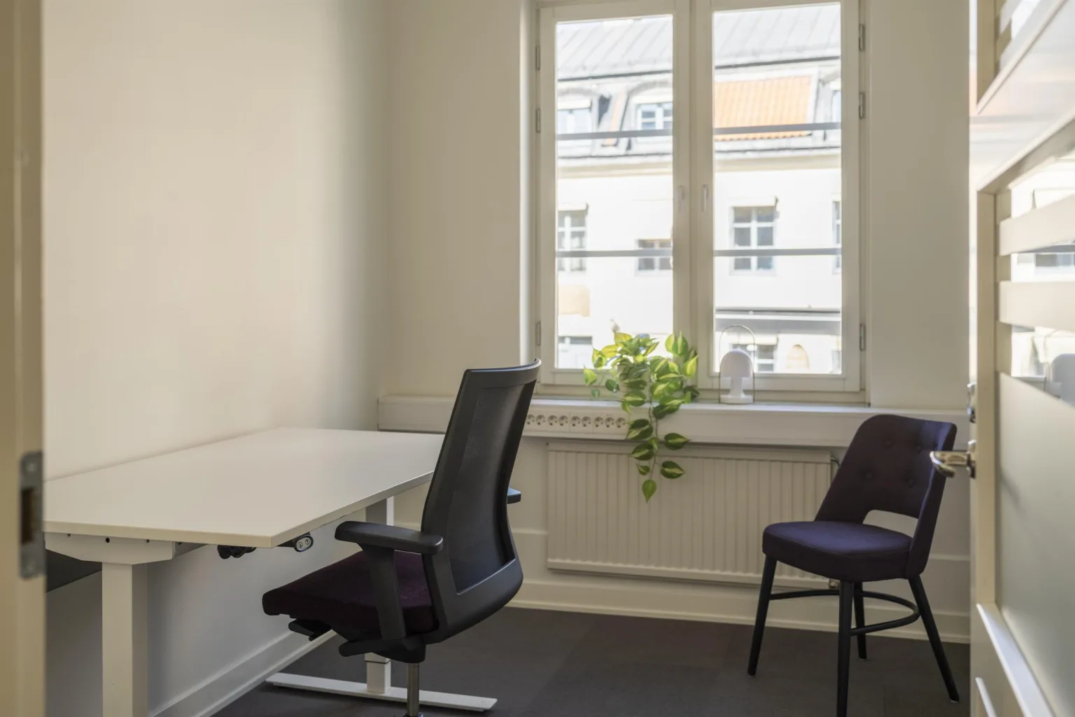 5. Litet kontorsrum med skrivbord, kontorsstol, extra stol och växt på fönsterbrädan.