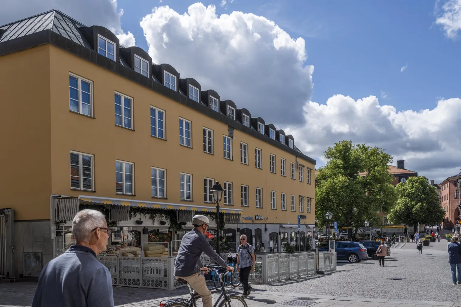 1. Gul byggnad med svarta takfönster och uteservering på trottoaren, med människor som promenerar och cyklar under en blå himmel med moln.