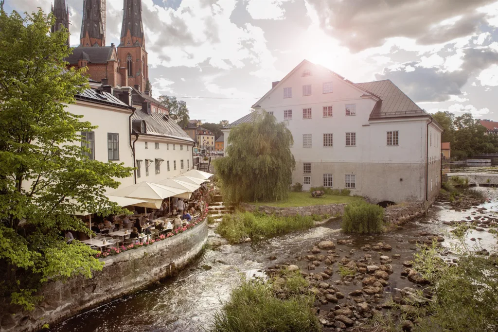 5. En utomhusscen med en å, uteserveringar, och en kyrka i bakgrunden under en solig himmel.