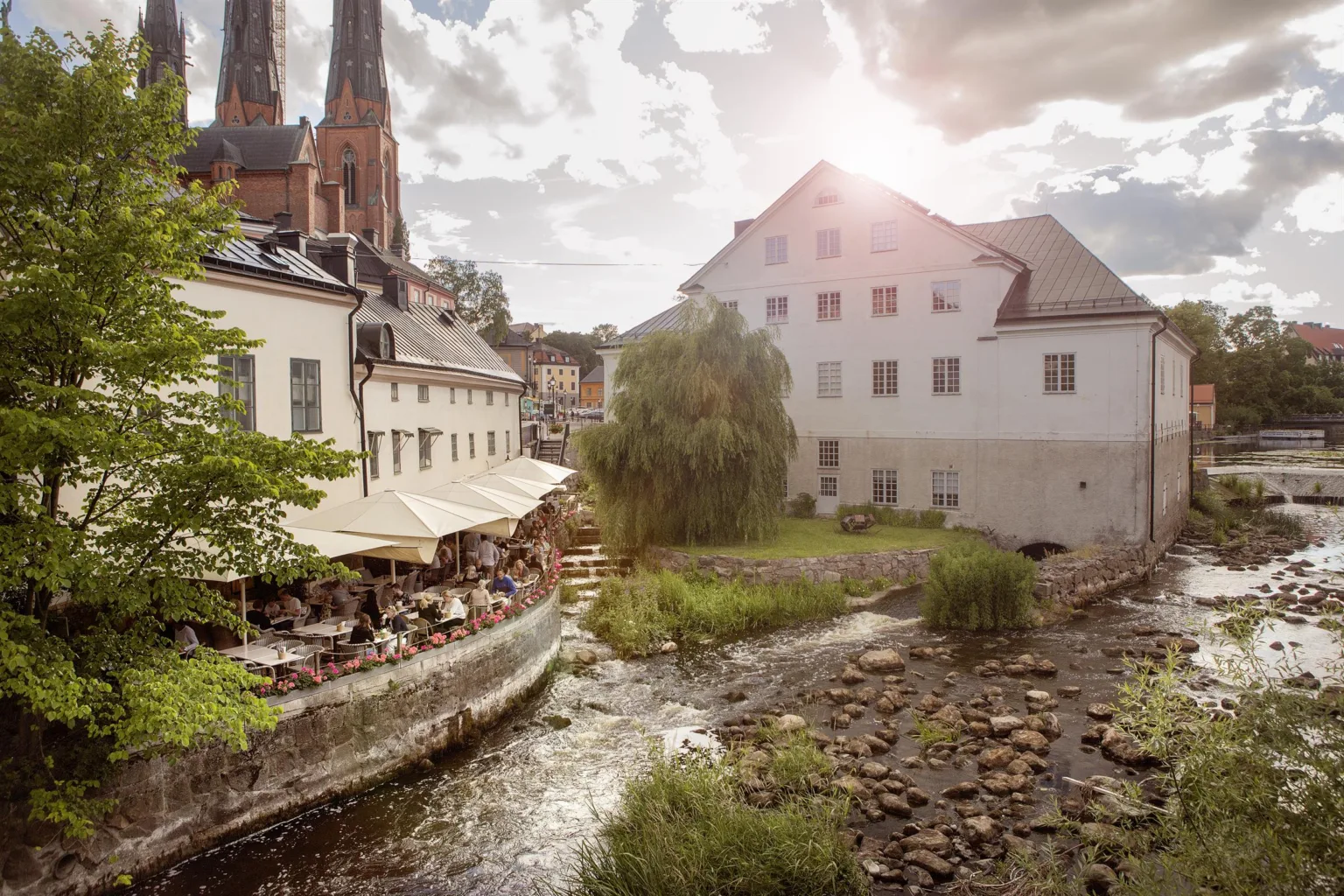 5. En utomhusscen med en å, uteserveringar, och en kyrka i bakgrunden under en solig himmel.