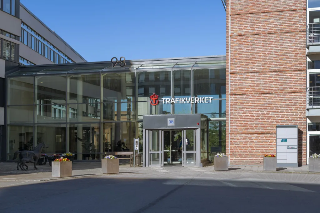 4. Ingången till en byggnad med glasfasad och texten "TRAFIKVERKET" ovanför dörren, omgiven av blomkrukor och en tegelvägg.