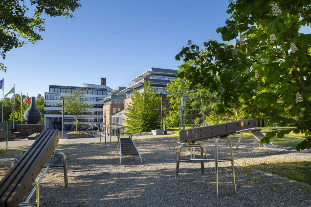 3. En utomhuslekplats med bänkar och lekutrustning, omgiven av träd och moderna byggnader i bakgrunden.