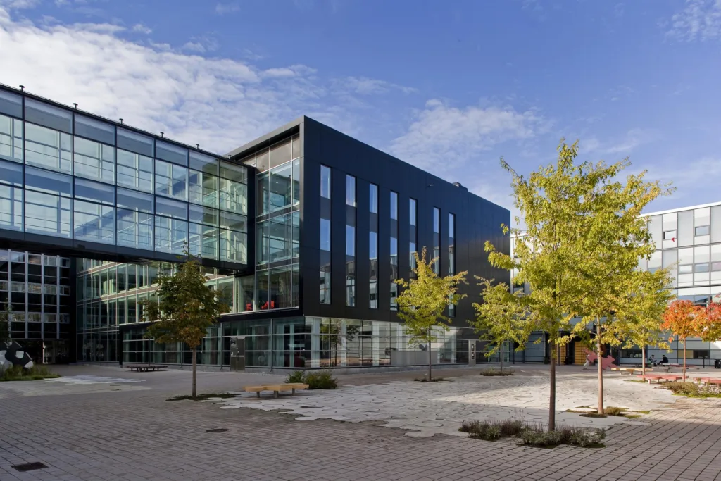 1. En modern byggnad med glasfasad och svarta paneler, omgiven av träd och en stenlagd innergård.