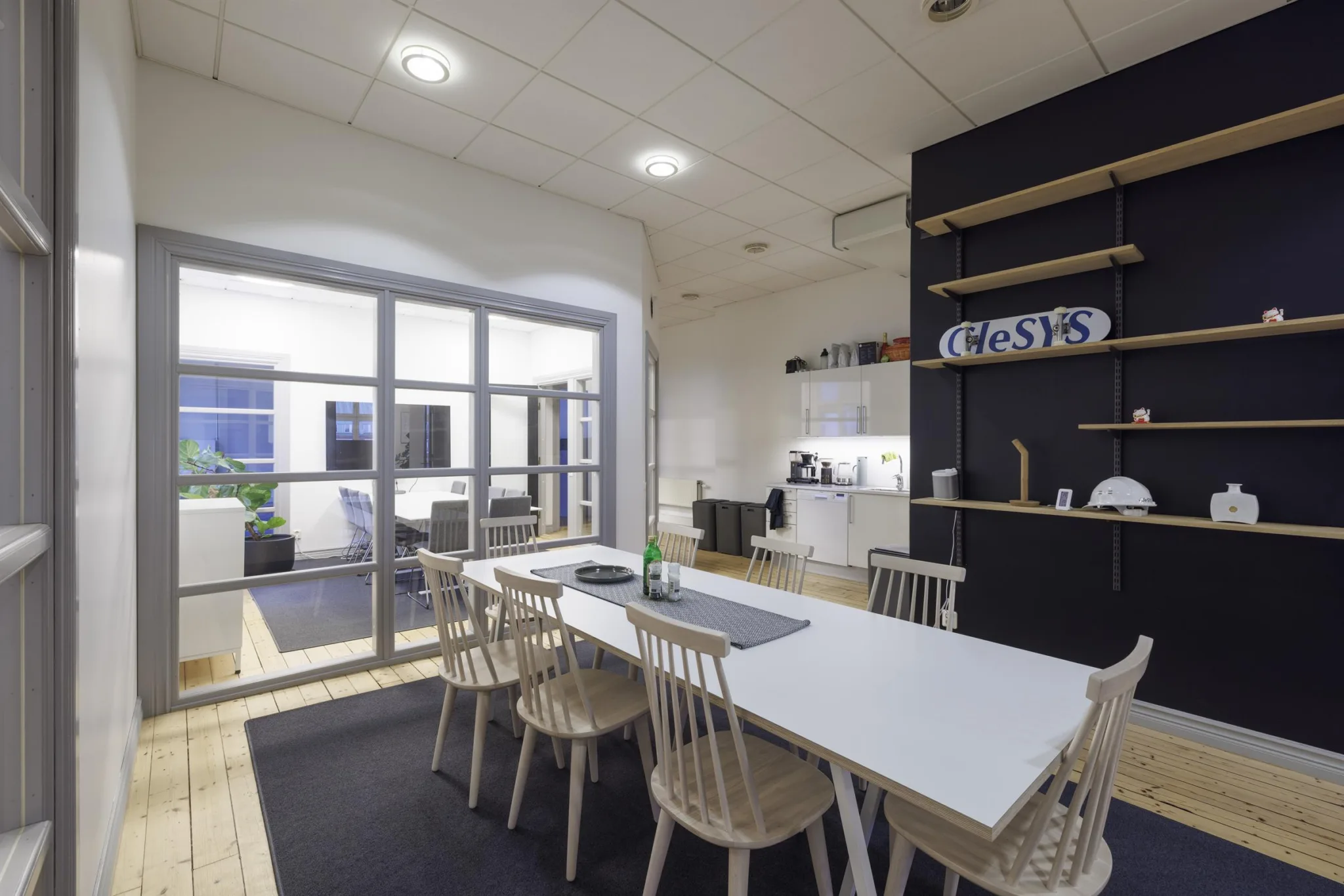 Ett mötesrum med ett långt bord, flera stolar och ett kök med kaffemaskin i bakgrunden.