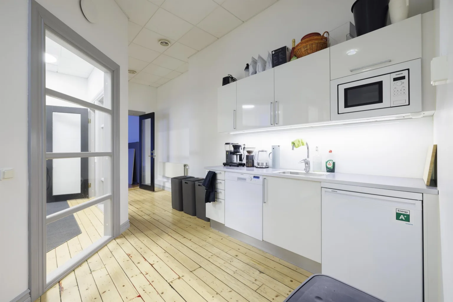 Ett kök med vitt kakel, vita skåp, mikrovågsugn och kaffemaskin, samt en öppning till en annan del av kontoret.