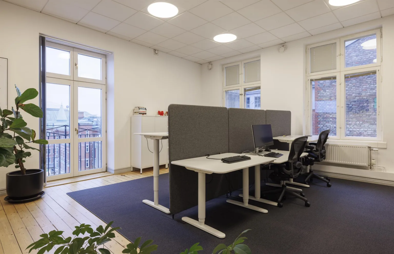 1. En modern arbetsplats med skrivbord, datorskärmar och stolar, omgiven av vita väggar och stora fönster.