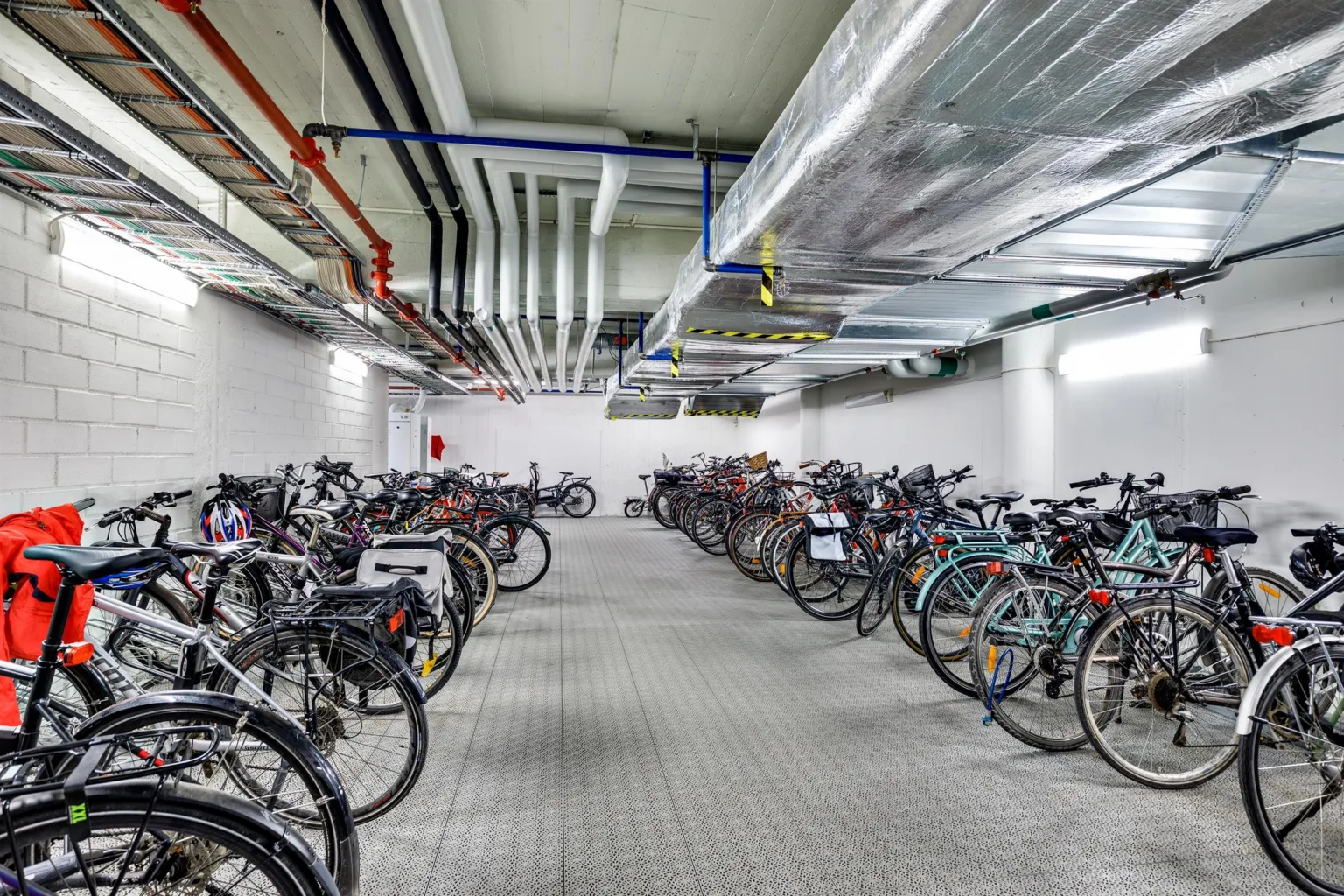 En cykelparkering inomhus med flera rader av cyklar och synliga rör i taket.