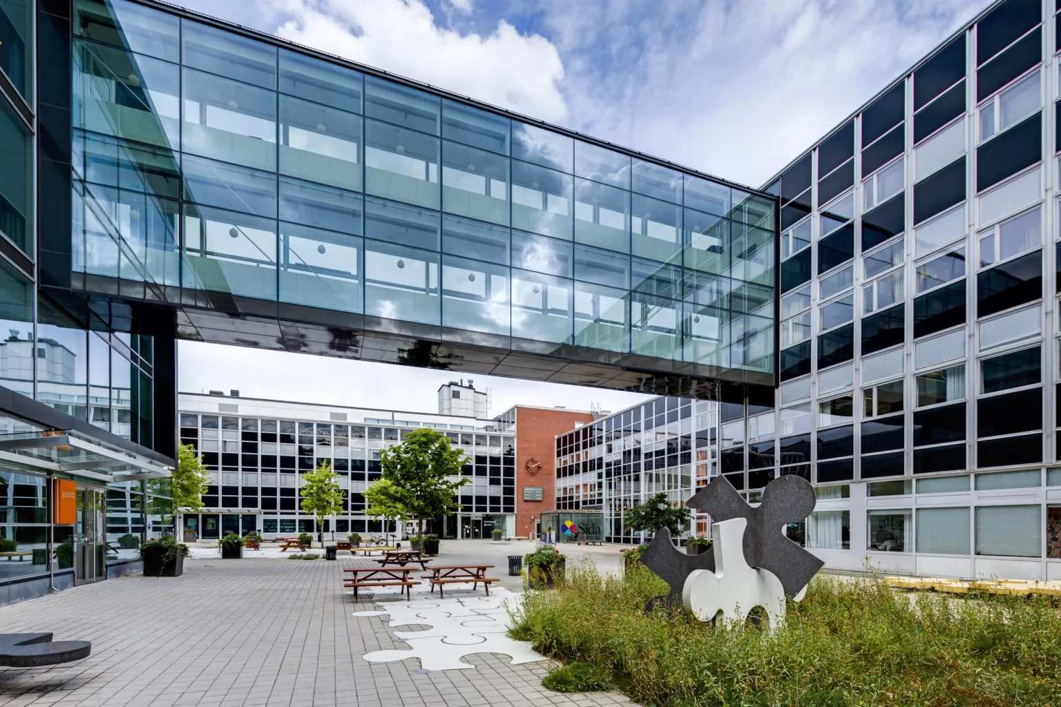 En modern byggnad med glasbro, omgiven av växter och utemöbler på en innergård.
