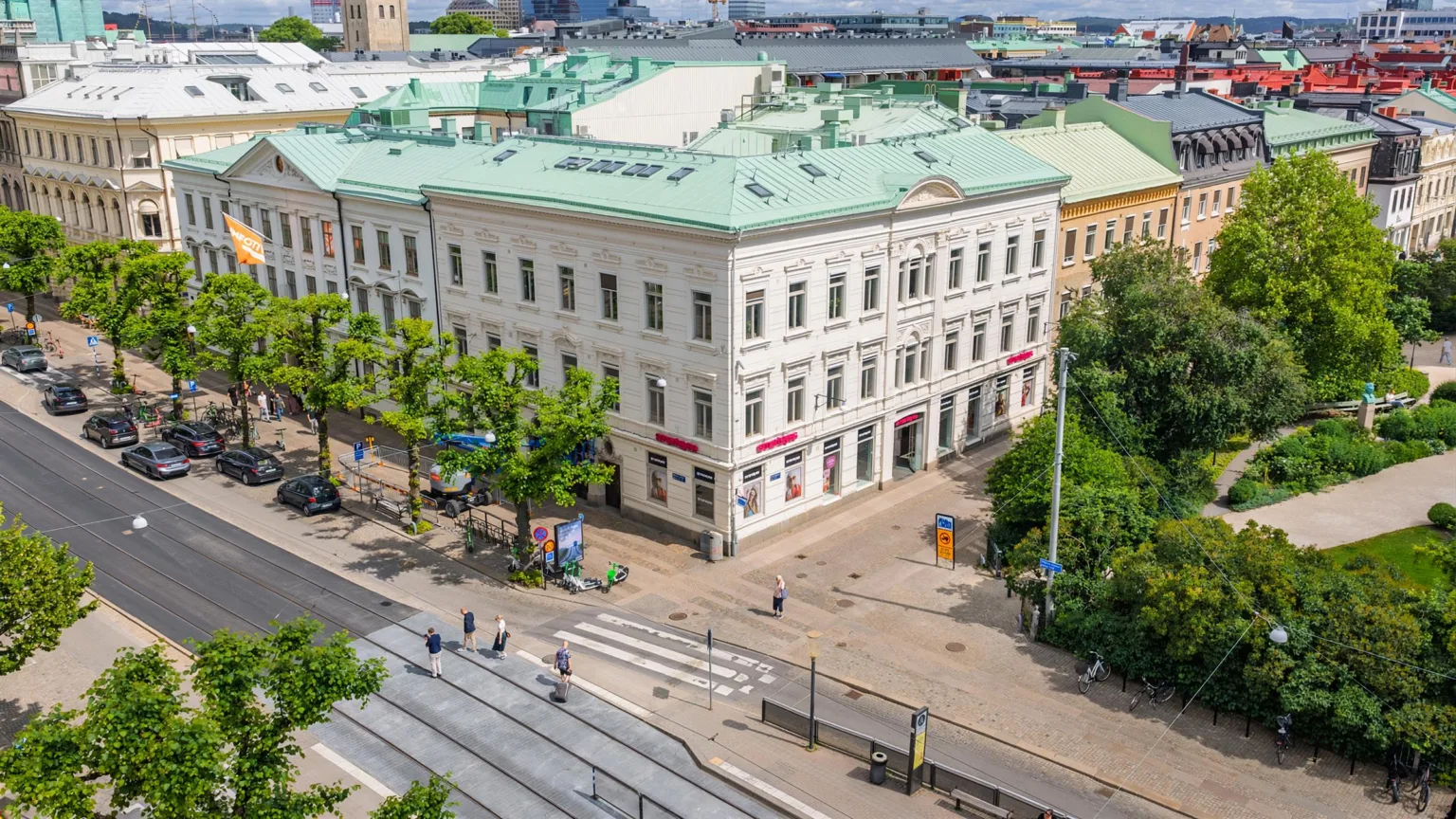 En vit byggnad med grönt tak, omgiven av träd och gångvägar, sedd från ovan.