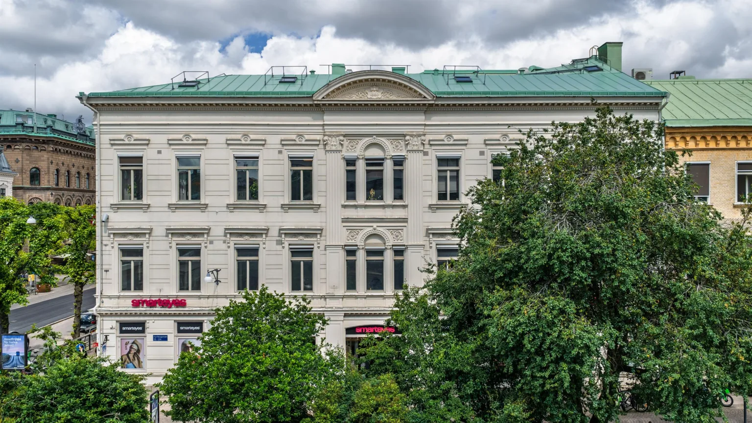 En vit byggnad med grönt tak, omgiven av grönskande träd, fotograferad under molnig himmel.