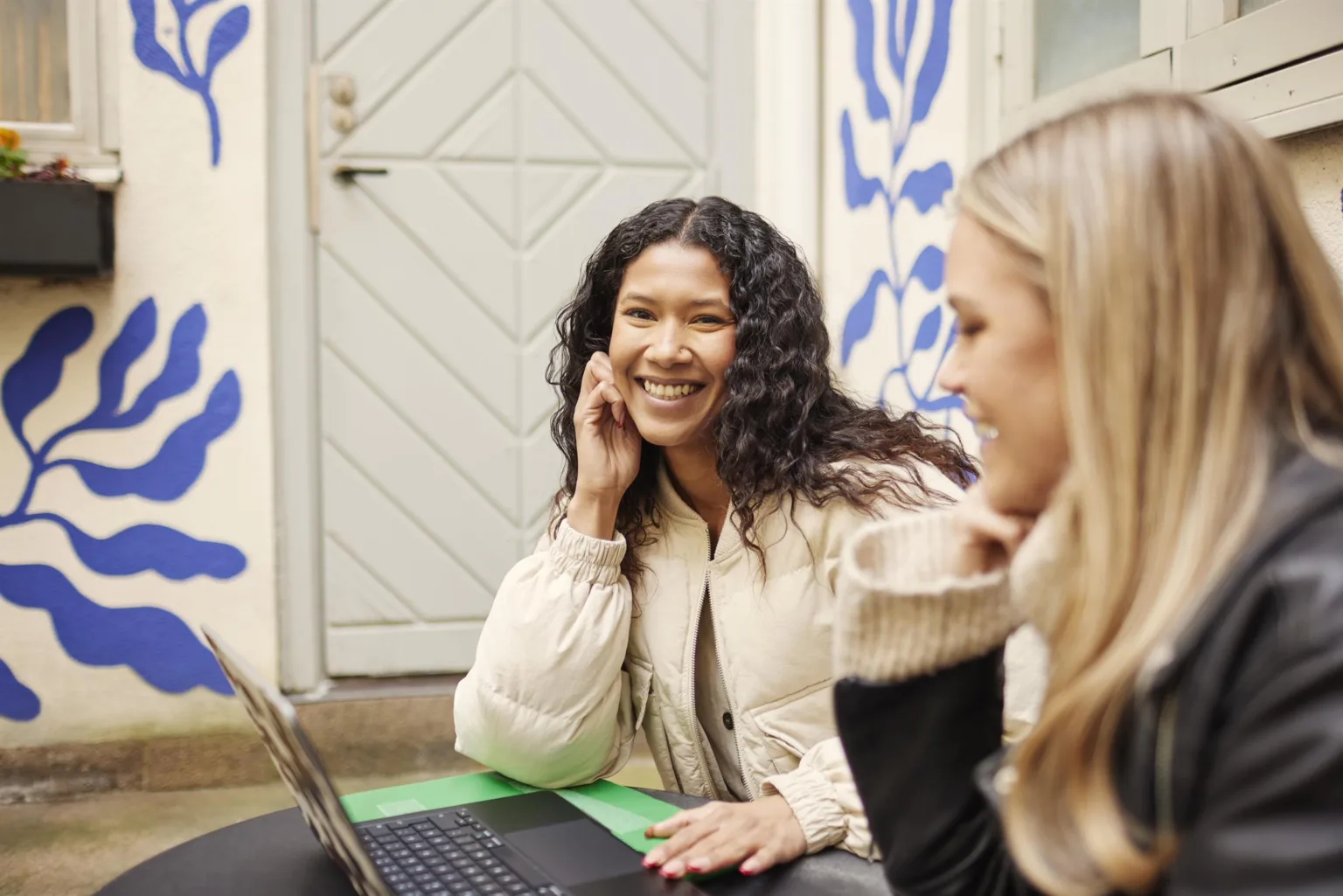 Två kvinnor sitter med en laptop och ler, med en vit dörr och blåmålade väggar i bakgrunden.