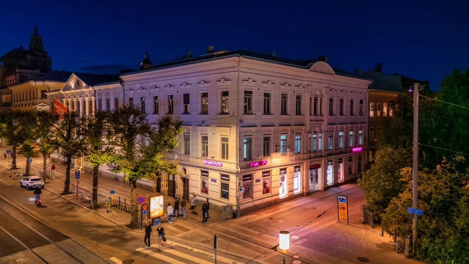 En vit byggnad med klassisk arkitektur, upplyst av gatlampor på kvällen. Fotgängare och trafik syns på gatan.