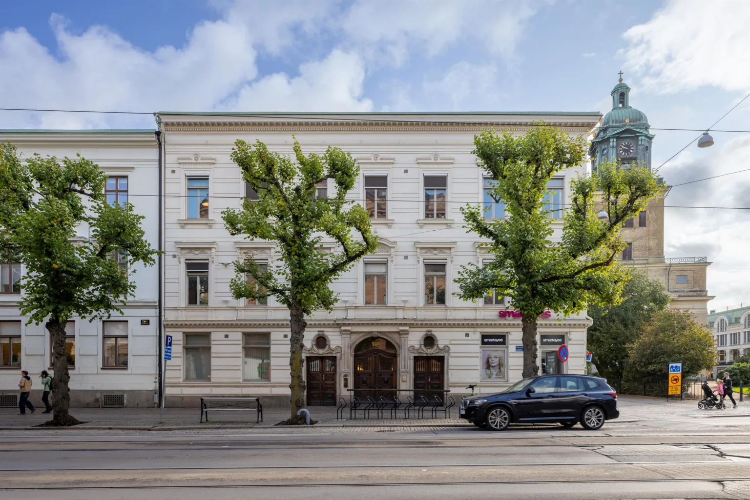 En vit byggnad med träd längs gatan, en parkerad bil och trottoar framför.