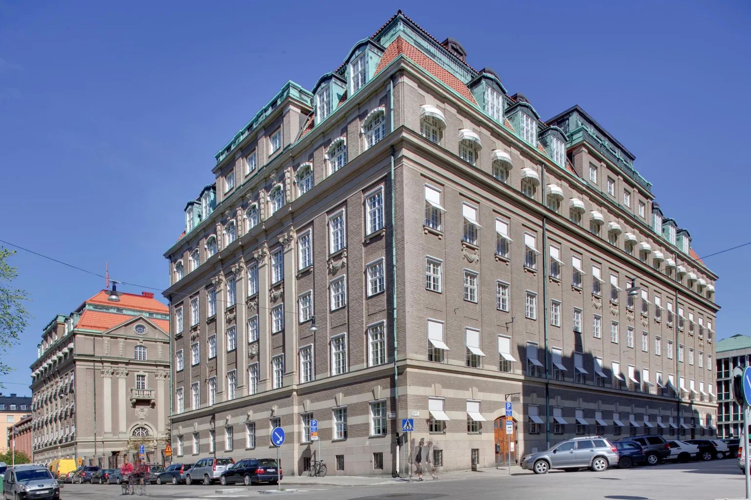 En stor byggnad med klassisk arkitektur, flera våningar och tak med gröna detaljer, belägen vid en gata med trottoarer.