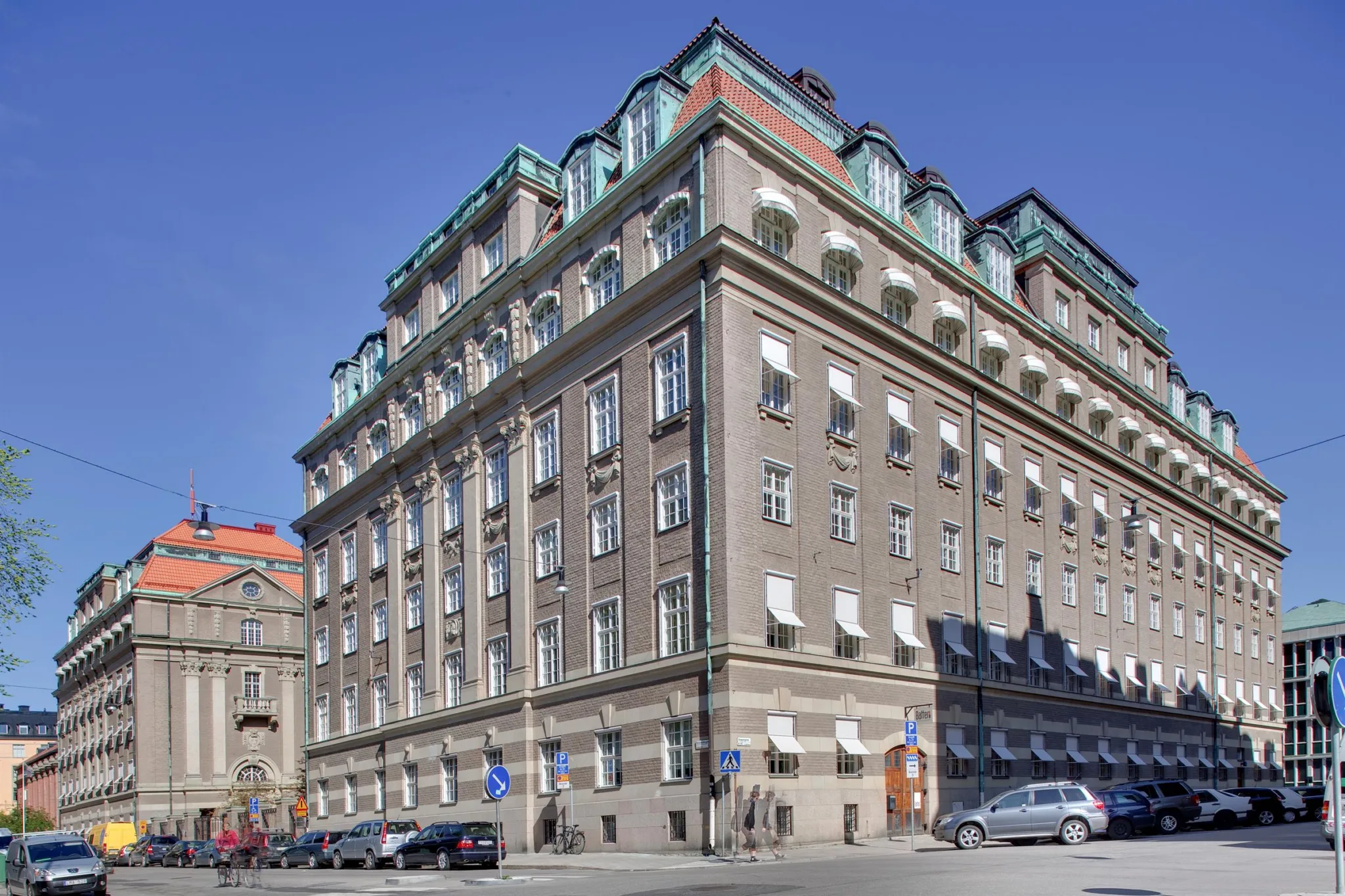 En stor byggnad med klassisk arkitektur, flera våningar och tak med gröna detaljer, belägen vid en gata med trottoarer.