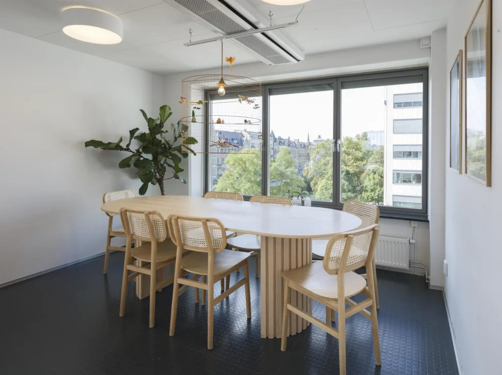 3. Ett konferensrum med ett runt bord, sex ljusa trästolar och fönster som vetter mot grönska och byggnader.