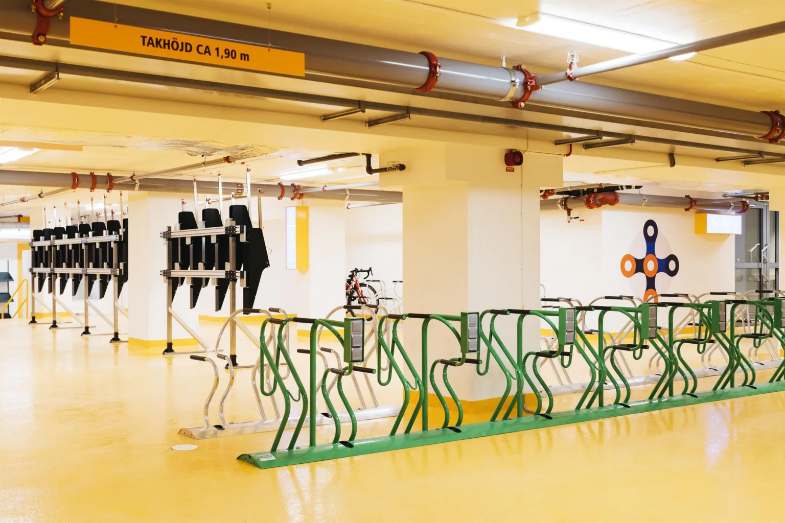 En cykelparkering inomhus med gröna cykelställ, gult golv och skylt med texten "Takhöjd ca 1,80 m".