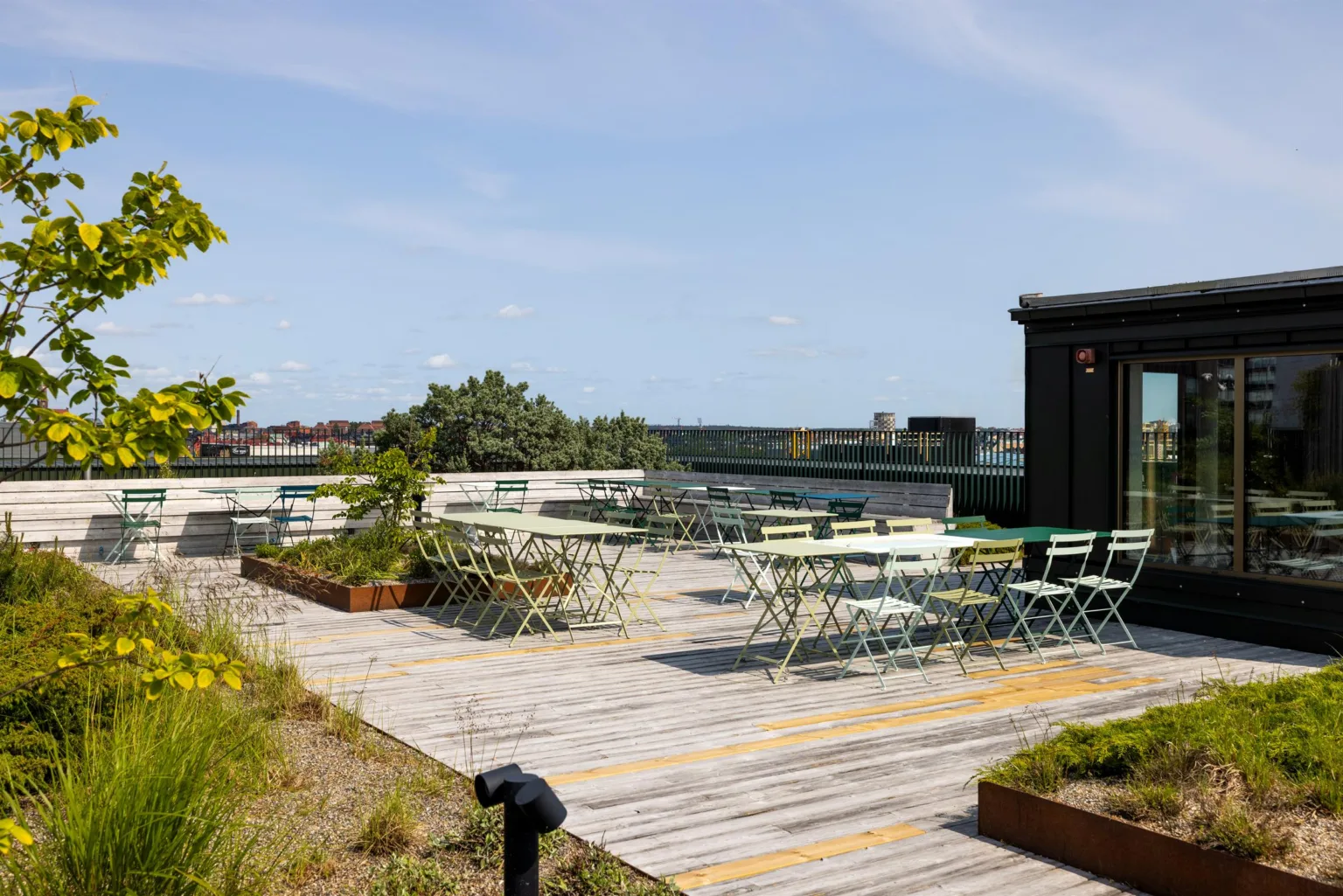 En takterrass med bord och stolar omgivna av grönska under en klarblå himmel.