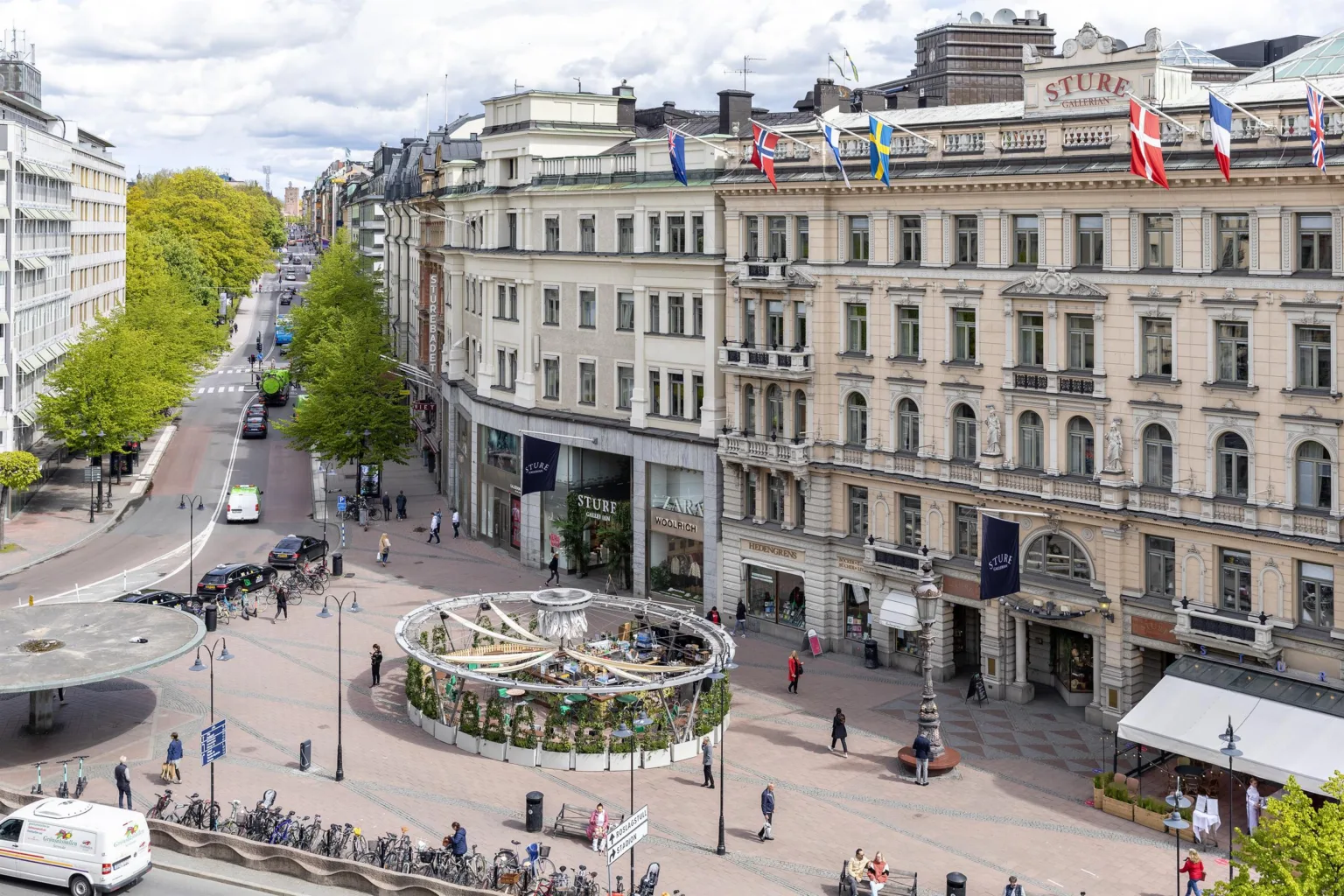 En vy över en stadsgata med stora byggnader, flaggor och en cirkulär planteringsyta omgiven av människor.